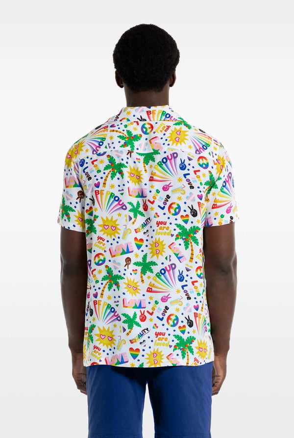 A man is wearing a white Hawaiian shirt with a rainbow pride print. The Print consists of peace symbols, hearts, and other icons. He is also wearing blue pants. He is smiling at the camera and holding the sides of the shirt. View from the back.