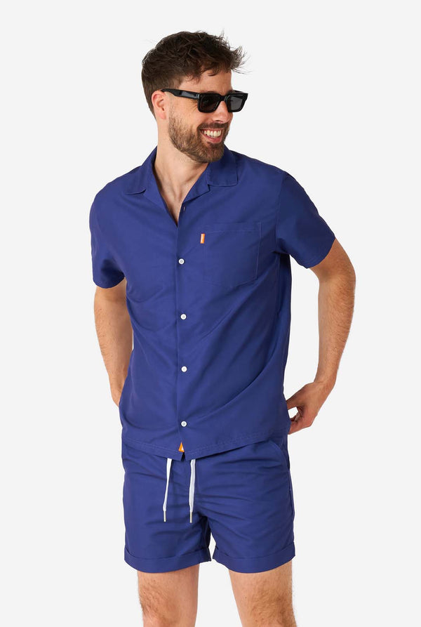 Man wearing blue summer set consisting of shirt and shorts and wearing sunglasses.