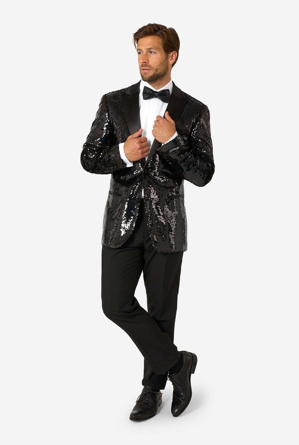 Full-body view of man in a black sequin tuxedo with satin lapels, standing with legs crossed and hands adjusting the jacket.