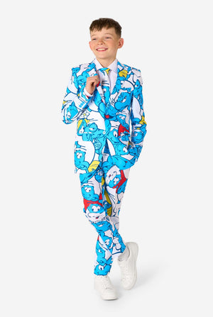Teen boy wearing a vibrant Smurfs kids suit with colorful cartoon character prints in blue, red, yellow, and white, paired with a matching tie and white shirt, smiling against a plain background. Full body with crossed legs.