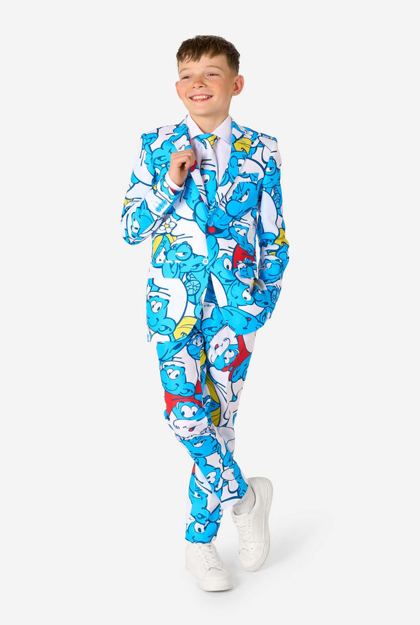 Teen boy wearing a vibrant Smurfs kids suit with colorful cartoon character prints in blue, red, yellow, and white, paired with a matching tie and white shirt, smiling against a plain background. Full body with crossed legs.