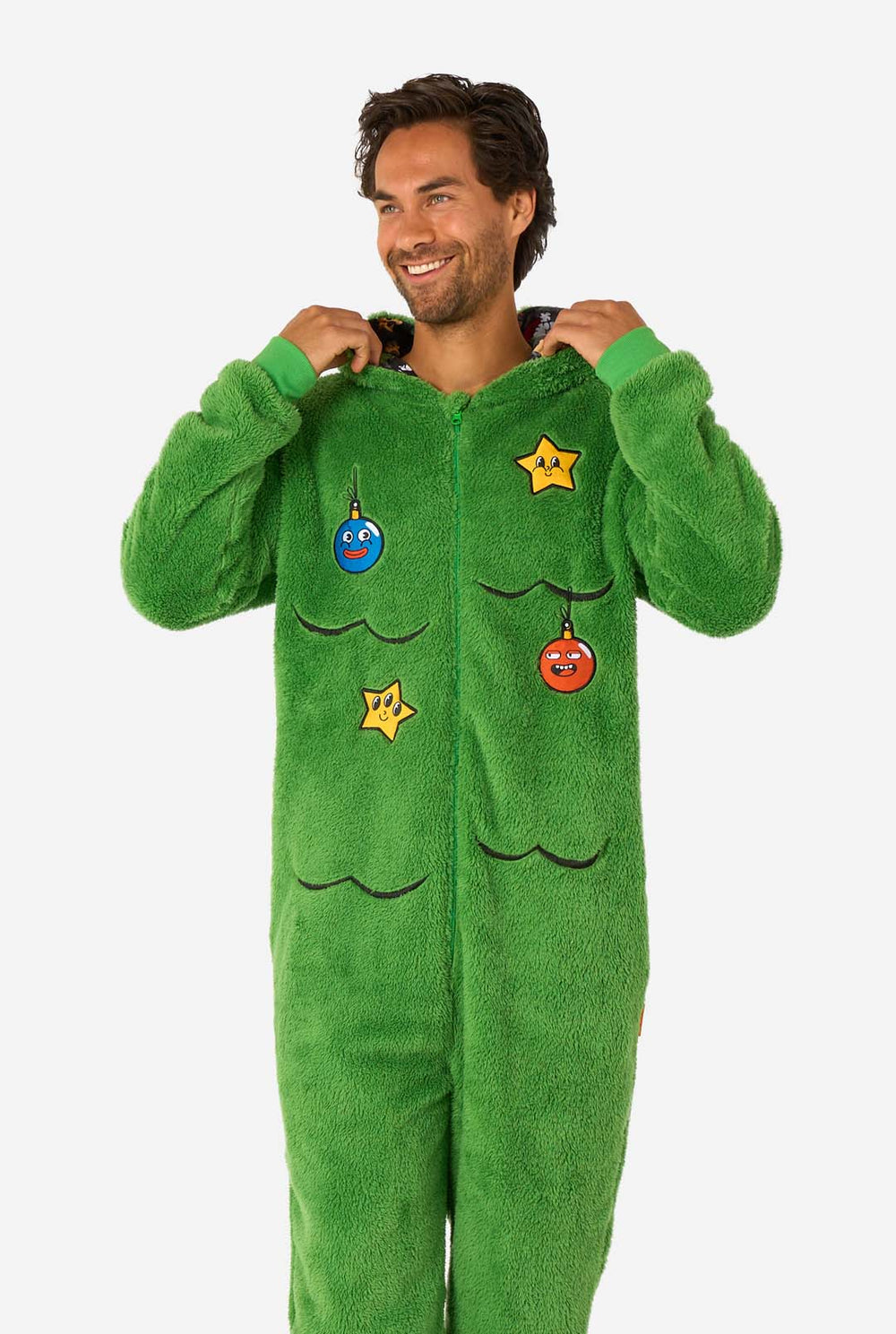 A smiling man wearing a green Christmas onesie designed to look like a Christmas tree, decorated with cartoon ornaments and stars. Hood on it's head