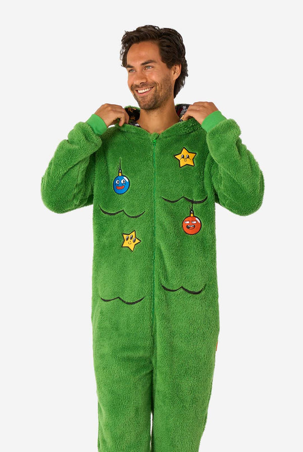 A smiling man wearing a green Christmas onesie designed to look like a Christmas tree, decorated with cartoon ornaments and stars.