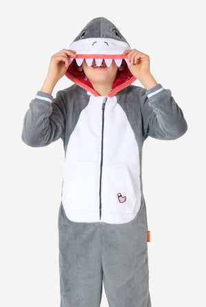 Child wearing a plush grey shark onesie with white belly panel, red inner hood, and attached shark eyes and teeth on the hood. The child lifts the hood’s mouth over their face while smiling.