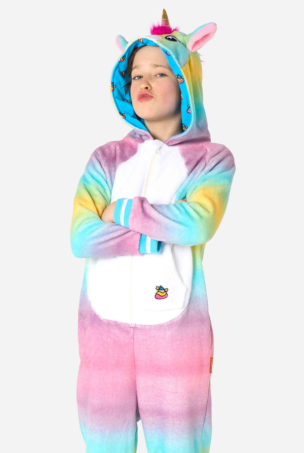 A smiling kid is wearing a white, pink, blue and yellow kids onesie designed as a unicorn. The Unicorn onesie features a hood resembling a unicorn head and other playfull details