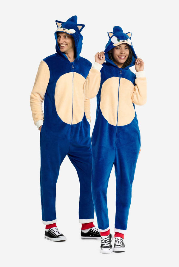 A man and woman wearing a playful blue and beige Sonic the Hedgehog-themed adult onesie from OppoSuits. The cozy onesie features a zip-up front and an attached hood designed to resemble Sonic’s iconic face, complete with ears and an expressive gaze. The blue body of the suit contrasts with a beige belly, mimicking Sonic's classic look. Full body photo.
