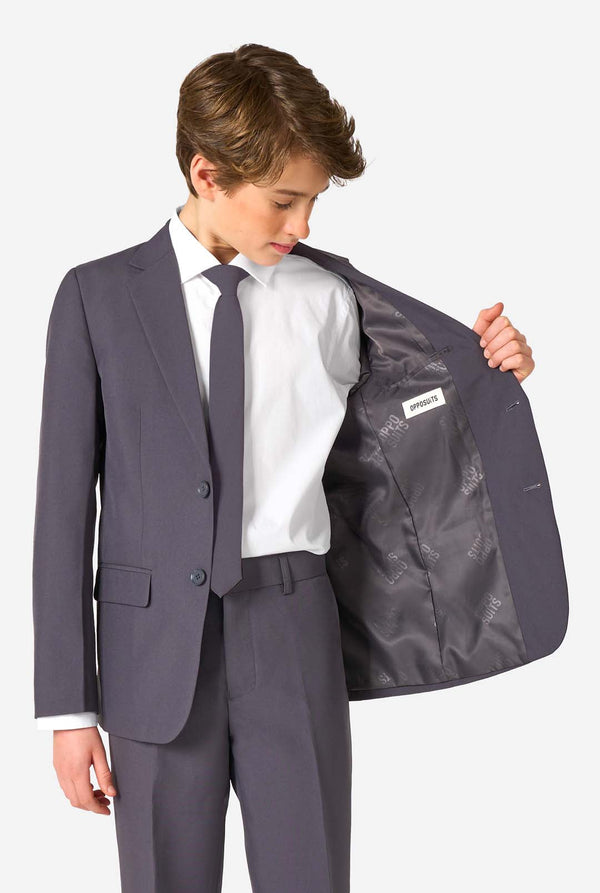 Teen boy wearing a stylish charcoal colored grey suit with a white dress shirt, smiling confidently. Perfect formal outfit for boys' special occasions, weddings, and parties. Holding one side of his jacket open.